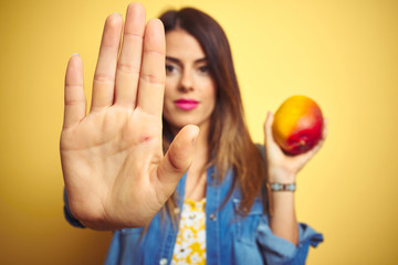 Young beautiful woman eating fresh healthy mango over yellow background with open hand doing stop...