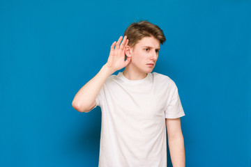 A young man in a white T-shirt holds his ear with an attentive look on a blue background. The man...