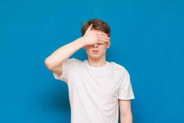 A mysterious man in a white T-shirt covers his face with his hands, against a blue background. Blue background.