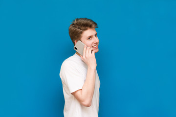 Portrait of a young man on a blue background holding a phone, happily smiling, looking at the camera, isolated on a blue background. An adult man in a white t-shirt rejoices.