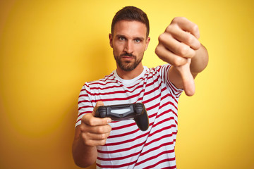 Young handsome man playing video games using joystick game pad over yellow background with angry face, negative sign showing dislike with thumbs down, rejection concept