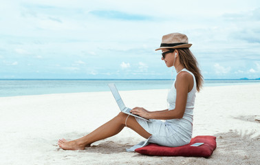 Young woman in a hat working with laptop computer on tropical island beach under palm trees...