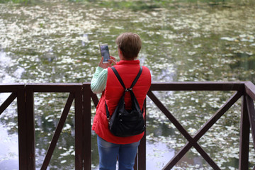 Fototapeta premium Mature woman with short hair in a red vest photographs a pond with duckweed using a smartphone