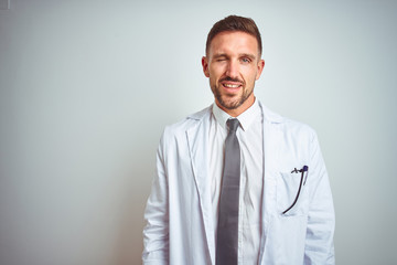 Young handsome doctor man wearing white profressional coat over isolated background winking looking at the camera with sexy expression, cheerful and happy face.