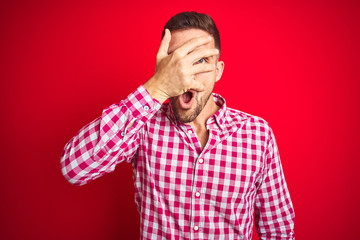 Fototapeta premium Young handsome man over red isolated background peeking in shock covering face and eyes with hand, looking through fingers with embarrassed expression.