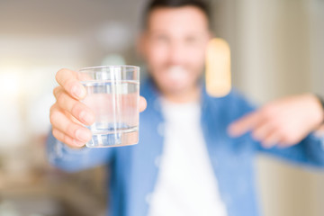 Young handsome man drinking a glass of water at home with surprise face pointing finger to himself