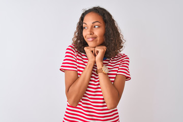 Young brazilian woman wearing red striped t-shirt standing over isolated white background laughing nervous and excited with hands on chin looking to the side