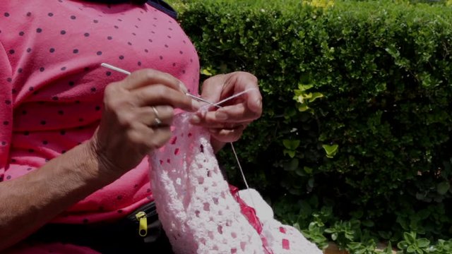 Detalle de las manos de una mujer anciana con blusa rosa tejiendo en el jard&iacute;n