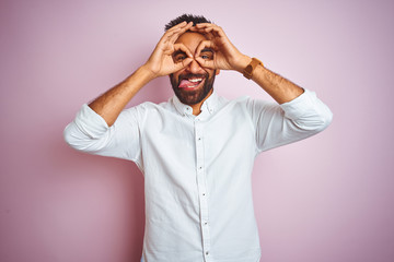 Young indian businessman wearing elegant shirt standing over isolated pink background doing ok gesture like binoculars sticking tongue out, eyes looking through fingers. Crazy expression.