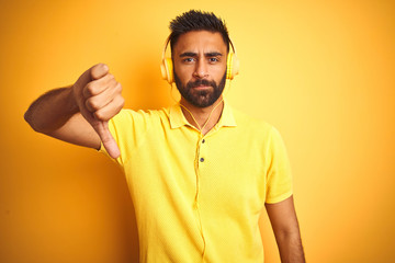 Arab indian hispanic man listening to music using headphones over isolated yellow background with angry face, negative sign showing dislike with thumbs down, rejection concept