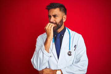 Young indian doctor man wearing stethoscope standing over isolated red background looking stressed and nervous with hands on mouth biting nails. Anxiety problem.