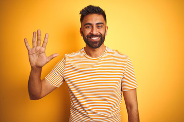 Young indian man wearing t-shirt standing over isolated yellow background showing and pointing up with fingers number five while smiling confident and happy.