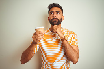 Young indian man drinking cup of coffee standing over isolated white background serious face...
