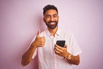 Young indian man using smartphone standing over isolated pink background happy with big smile doing...