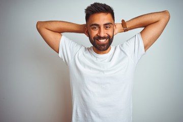 Young indian man wearing t-shirt standing over isolated white background relaxing and stretching, arms and hands behind head and neck smiling happy