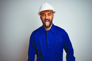 Handsome indian worker man wearing uniform and helmet over isolated white background sticking tongue out happy with funny expression. Emotion concept.