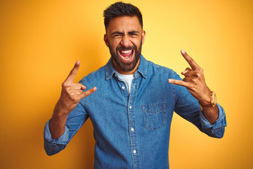 Young indian man wearing denim shirt standing over isolated yellow background shouting with crazy...