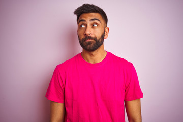 Young indian man wearing t-shirt standing over isolated pink background smiling looking to the side...