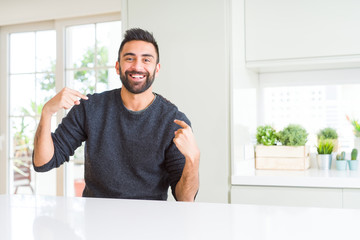 Handsome hispanic man wearing casual sweater at home looking confident with smile on face, pointing...