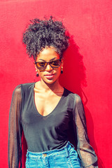 Portrait of Young African American Woman in New York City. Young black woman with afro hairstyle wearing long sleeve mesh sheer shirt, sunglasses, standing against red background under sun..