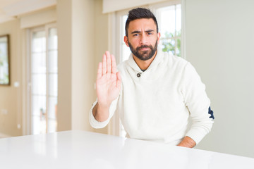 Handsome hispanic man wearing casual white sweater at home doing stop sing with palm of the hand. Warning expression with negative and serious gesture on the face.