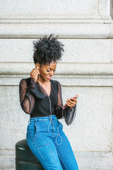 Young African American Woman with afro hairstyle, wearing mesh sheer long sleeve shirt blouse, blue jeans, sitting on street in New York City, listening music with earphone and cell phone, texting..