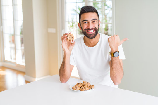 Handsome hispanic man eating chocolate chips cookies pointing and showing with thumb up to the side with happy face smiling
