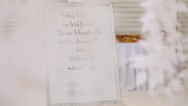 frame with invitation notice to wedding party for guests on table and white leaves in decorated banquet hall closeup