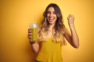 Young beautiful woman drinking detox green smoothy over yellow isolated background screaming proud and celebrating victory and success very excited, cheering emotion