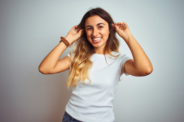 Fototapeta premium Young beautiful woman wearing casual white t-shirt over isolated background Smiling pulling ears with fingers, funny gesture. Audition problem