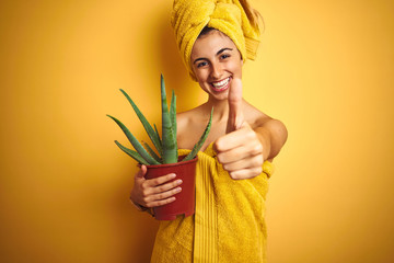 Young beautiful woman wearing a towel holding cactus pot over yellow isolated background happy with big smile doing ok sign, thumb up with fingers, excellent sign