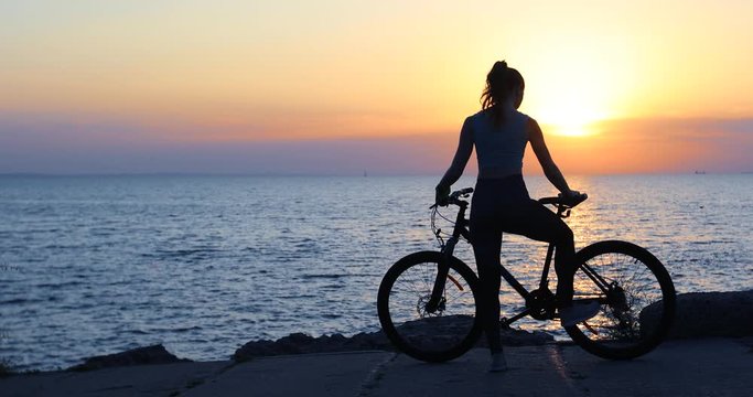sporty woman with bicycle walks along sea shore stops and prepares for evening workout against sunset backside view
