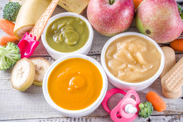 Infant baby food. Bowls with vegetable fruit puree, green, orange, yellow colors - broccoli, carrots, banana, apple. With baby accessories and toys wooden white background Flat lay top view