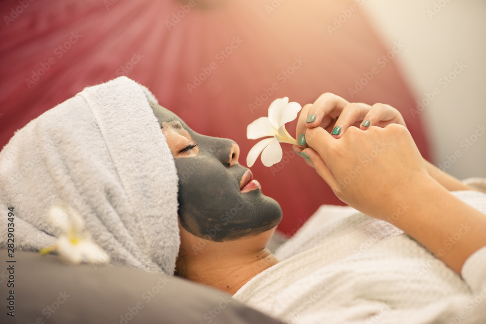 Wall mural young asian woman beautiful in clay mask she is smell a flower. woman spa face mask in spa uniform.