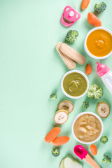 Fototapeta na wymiar Infant baby food. Bowls with vegetable fruit puree, green, orange, yellow colors - broccoli, carrots, banana, apple. With baby accessories and toys green background Flat lay top view copy space