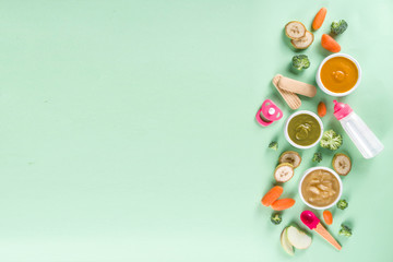 Infant baby food. Bowls with vegetable fruit puree, green, orange, yellow colors - broccoli, carrots, banana, apple. With baby accessories and toys green background Flat lay top view copy space