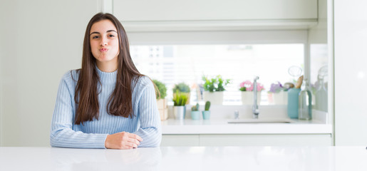 Wide angle picture of beautiful young woman sitting on white table at home puffing cheeks with funny face. Mouth inflated with air, crazy expression.