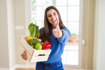 Beautiful young woman holding wooden box full of healthy groceries happy with big smile doing ok sign, thumb up with fingers, excellent sign