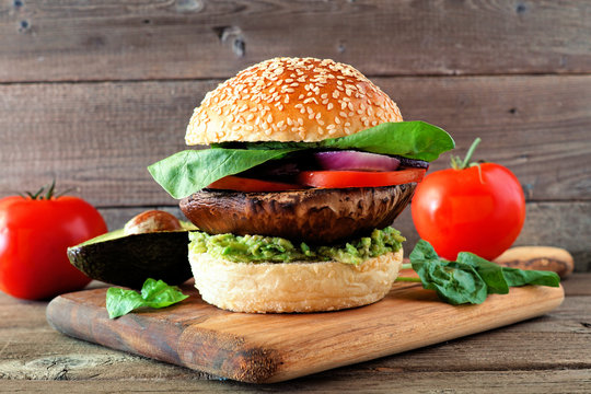 Portobello mushroom vegan burger with avocado, tomato, spinach and onion on a wooden serving board against a dark wood background