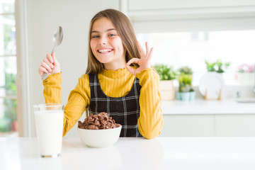 Beautiful young girl kid eating chocolate cereals and glass of milk for breakfast doing ok sign...