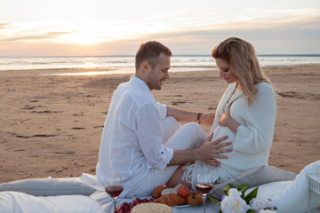Fototapeta premium Picnic on the beach. A man and pregnant woman had a picnic. A woman holds a peach in her hand, a man touches her tummy.
