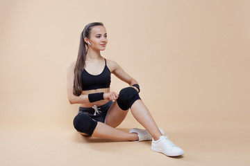 A young brunette girl with a calm smile is preparing for a workout, tying up her knee pads and bandages on her hands.