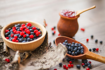 Blueberry and raspberry berry jam in a clay pot, a wooden spoon with fresh berries and birch bark on a wooden background