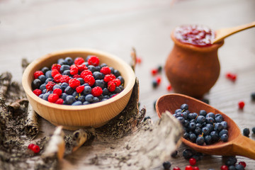Blueberry and raspberry berry jam in a clay pot, a wooden spoon with fresh berries and birch bark on a wooden background