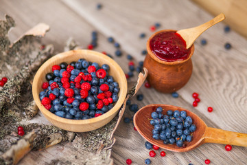 Blueberry and raspberry berry jam in a clay pot, a wooden spoon with fresh berries and birch bark on a wooden background