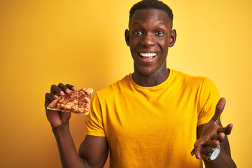 African american man eating slice of pizza standing over isolated yellow background very happy and excited, winner expression celebrating victory screaming with big smile and raised hands
