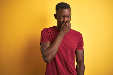 Young african american man wearing red t-shirt standing over isolated yellow background smelling something stinky and disgusting, intolerable smell, holding breath with fingers on nose. Bad smells 