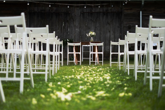 Wooden barn decorated by white cloth and flowers with greenery standing in the center of wedding ceremony. white chairs on side.