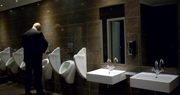 Bald man businessman in suit in between work pees in urinal in public toilet