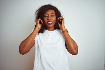 Young african american woman wearing t-shirt standing over isolated white background covering ears with fingers with annoyed expression for the noise of loud music. Deaf concept.